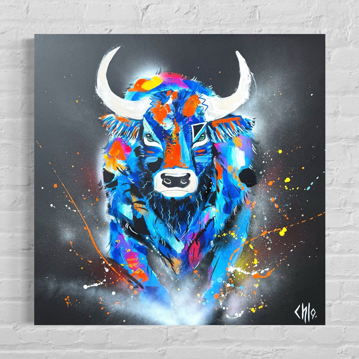 Peinture contemporaine colorée représentant un taureau de face, aux cornes blanches, réalisée dans un style abstrait et expressif, avec des éclaboussures de peinture bleue, orange, rose et jaune sur fond sombre, symbolisant la force et la puissance.